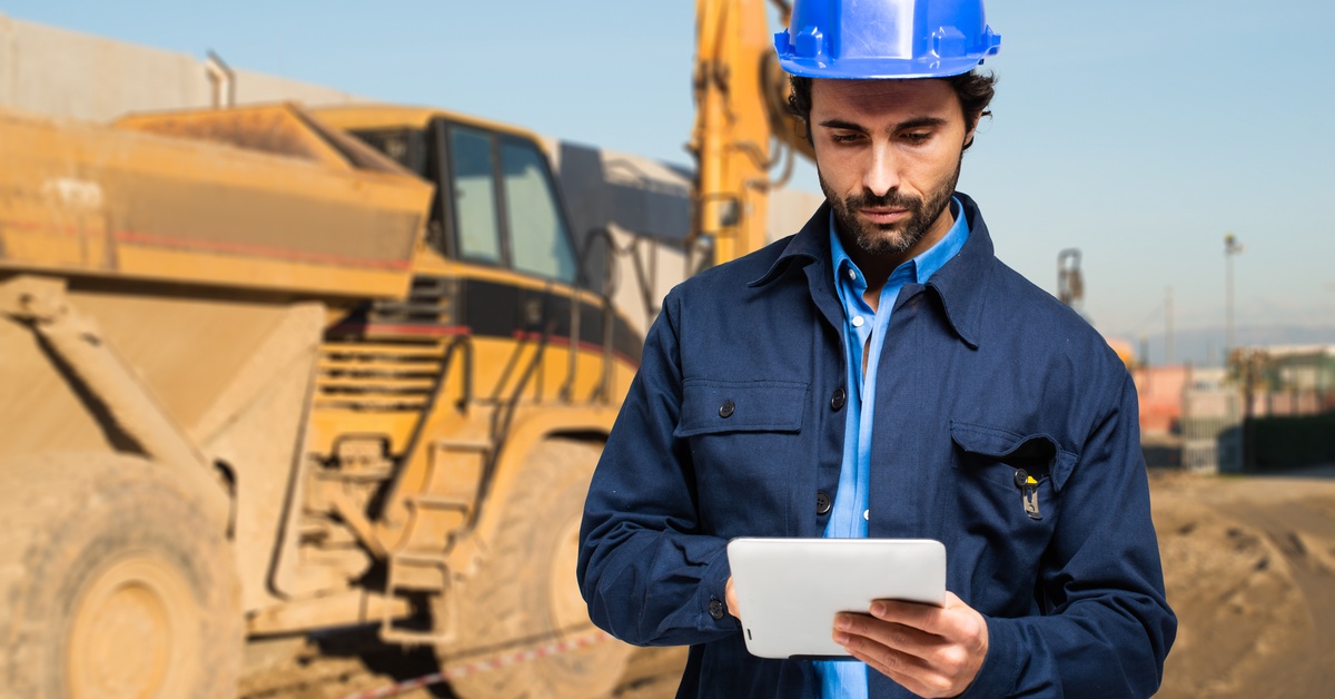 A construction worker in a hard hat uses a tablet computer at a construction site, with heavy equipment behind him.