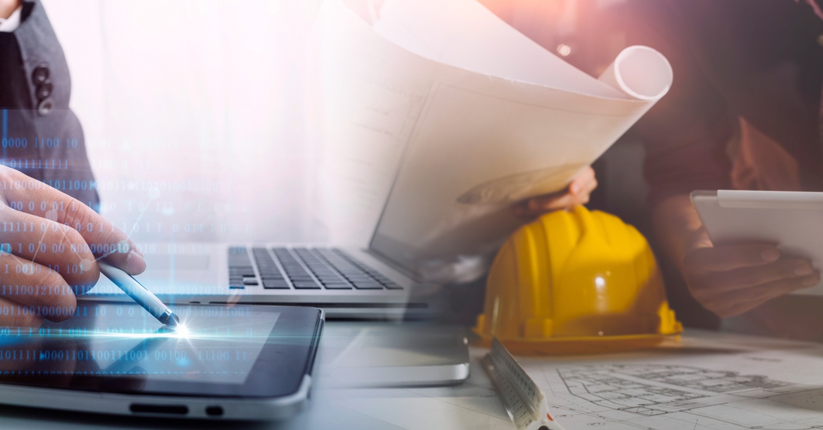 Two colleagues sit at a desk and work on a tablet and a laptop. There’s also a yellow hard hat on the desk.