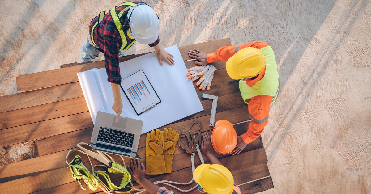 An aerial view of engineers and architects in safety gear discussing blueprints and data on a construction site.