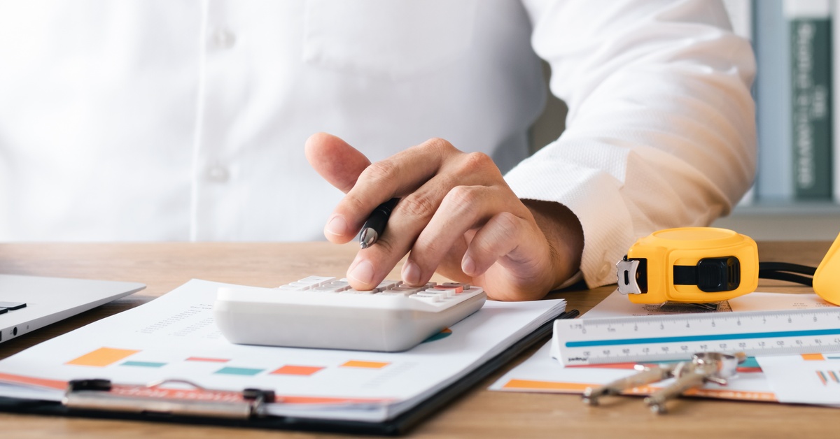 A partly seen businessman working at a desk with a laptop, calculator, documents, and a yellow hard hat.