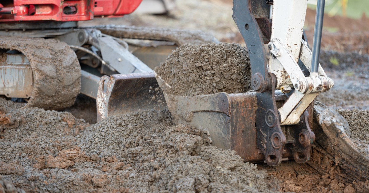 An excavator scoops earth with its bucket at a construction site, with dirt and gravel visible in the foreground.