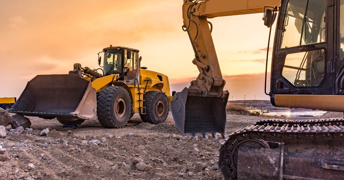 A group of construction vehicles, including an excavator and a wheel loader, work on a site under a warm, glowing sky.