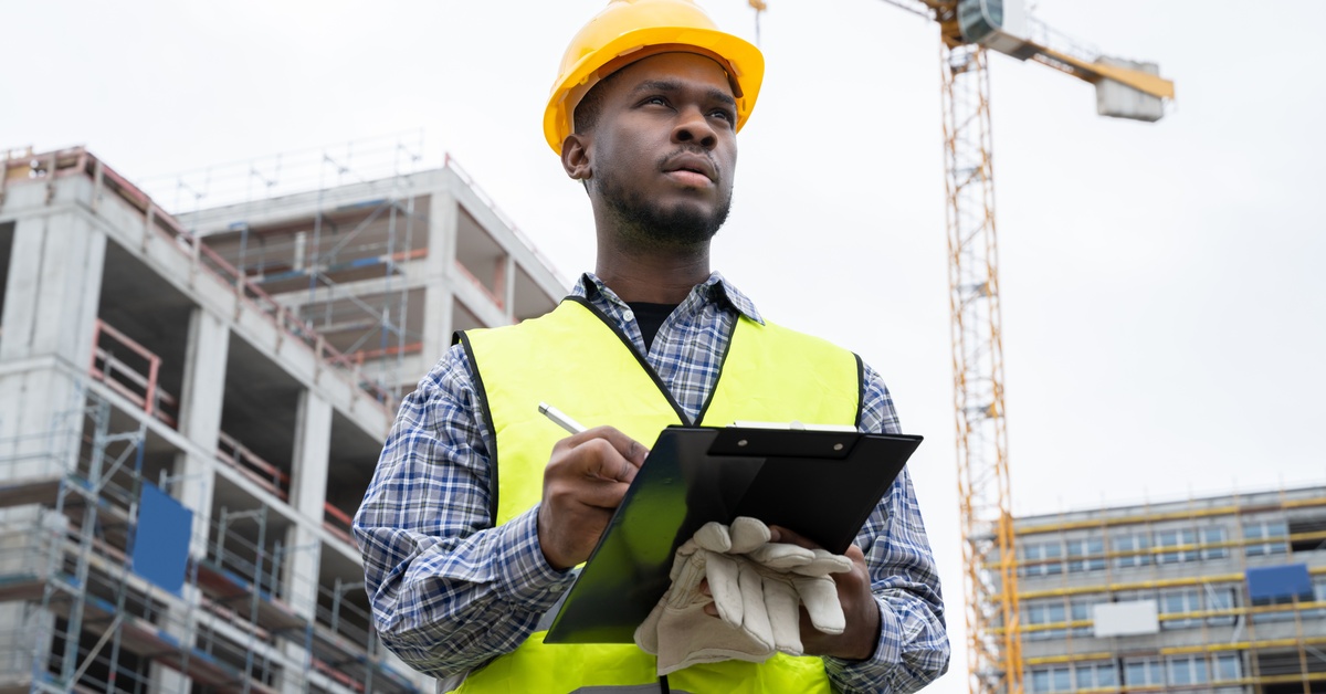 A man at a construction site wears a reflective safety vest and hard hat while holding a black clipboard