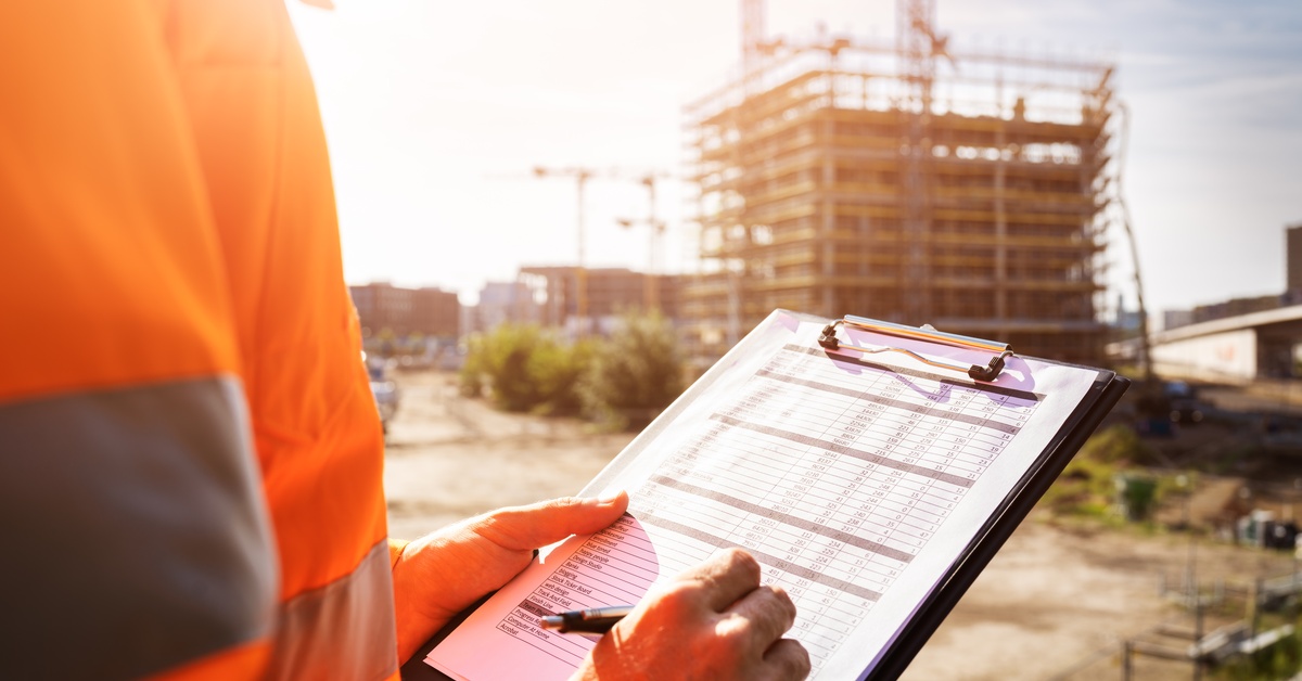 A partially seen construction worker at a job site; the worker wears an orange reflective vest and holds a clipboard.