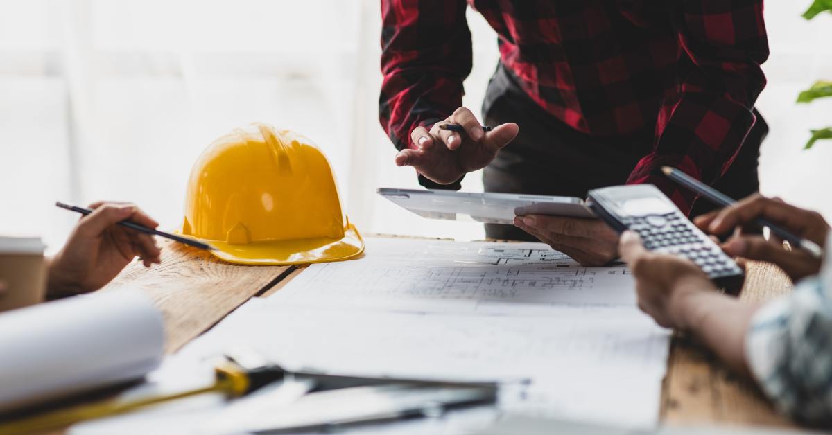 People review papers and a tablet on a wooden table with blueprints, a calculator, and a yellow hard hat.