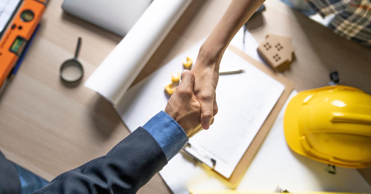 Two partly seen people shake hands across a desk with blueprints, a yellow hard hat, and building tools nearby.