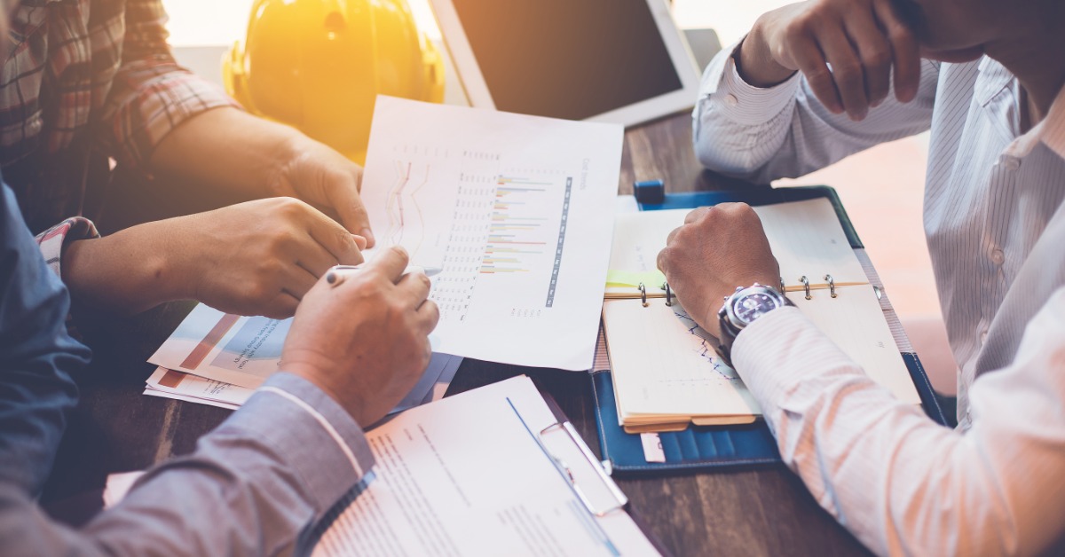Three employees sit around a desk with paperwork and a tablet; a hard hat rests on it; one worker points to a paper.