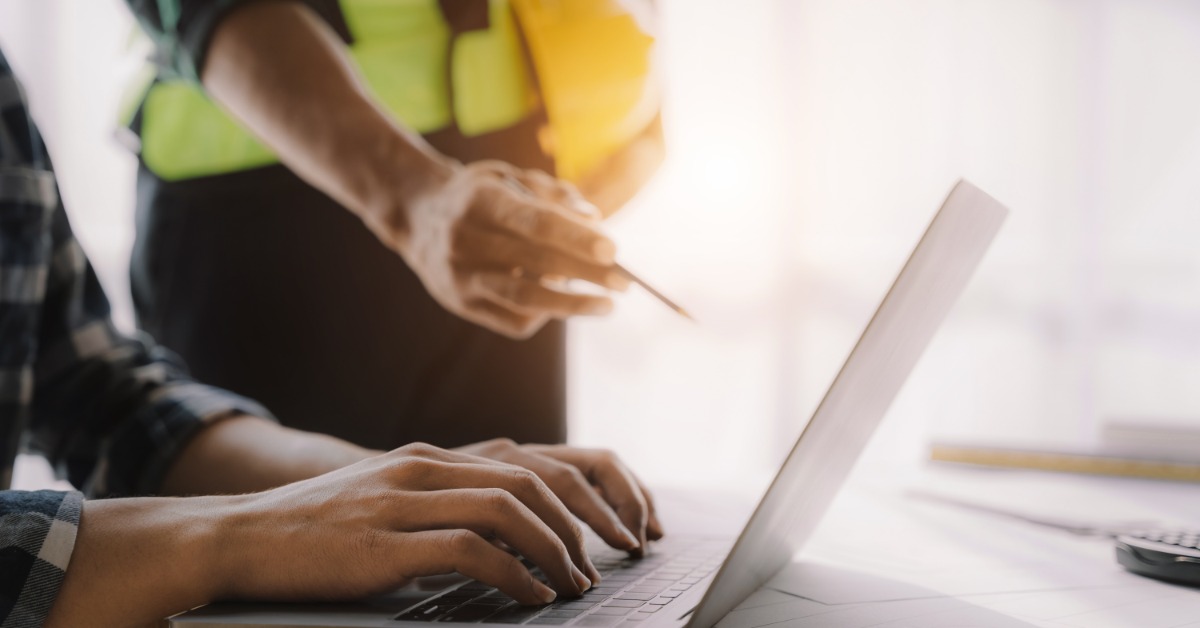 A person types on a laptop as someone wearing a yellow high-visibility vest and holding a hard hat points at the laptop.