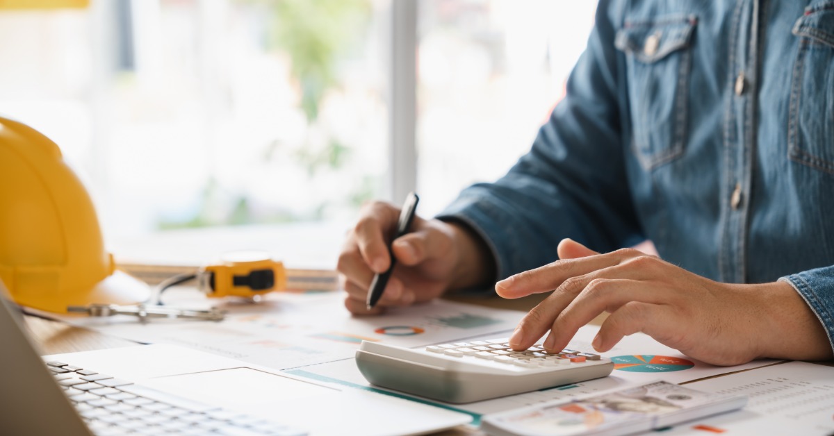 A person uses a calculator and pen while looking at financial documents at a desk with a laptop and a hard hat.