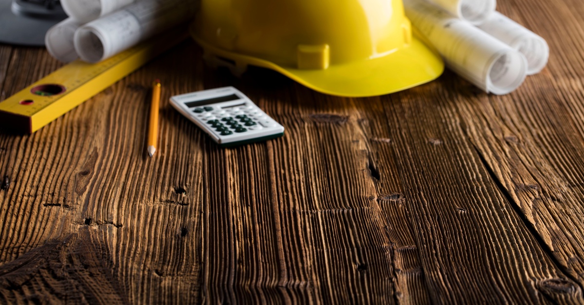 A yellow hard hat, rolled blueprints, a calculator, a pencil, and a measuring tool sit on a wooden surface.