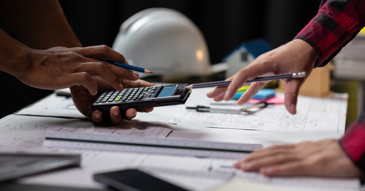 Hands use a calculator on a desk, with printed blueprints, tools, and papers spread across the surface.