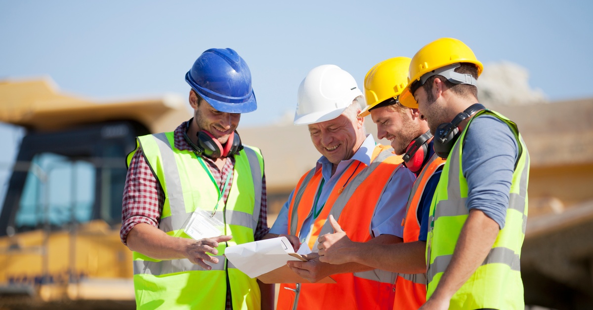 Four workers wearing hard hats and safety vests stand together looking at a document at an outdoor worksite.