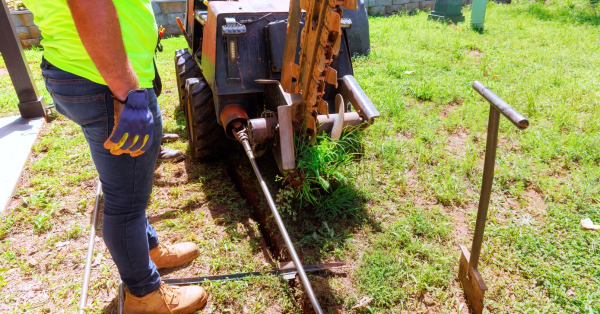 A worker stands beside a horizontal drilling machine near a ground opening with a pipe and tools positioned along the edge.