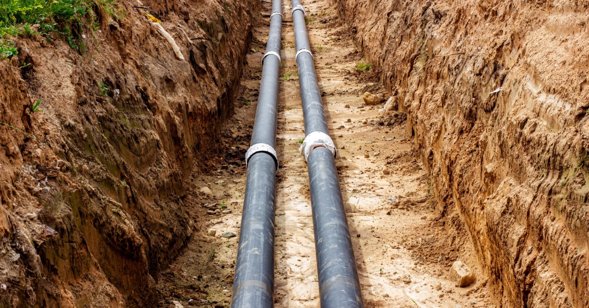 Two pipes run through a long, narrow trench in the ground, with exposed soil walls visible on both sides.