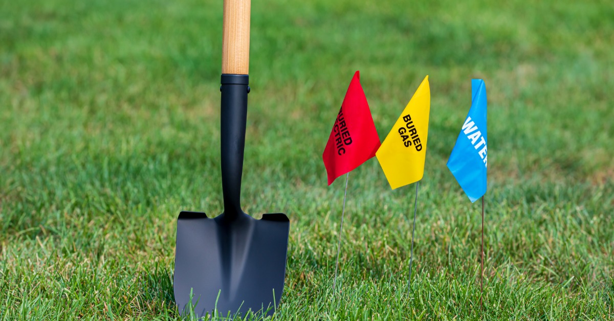 A shovel sits in the grass beside three small colored flags marking underground utility lines in an outdoor setting.