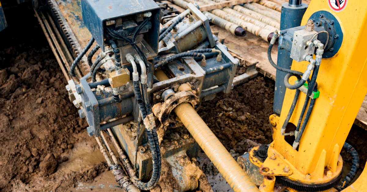 A close-up of a drilling machine guiding a pipe through muddy ground, with cables and metal rods around it.