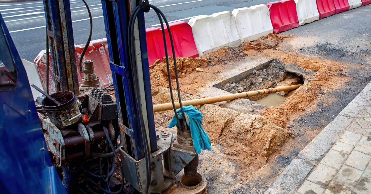 A drilling machine beside a roadside trench, with pipes and barriers near a paved sidewalk and road.