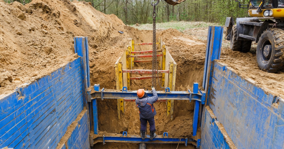 A worker standing in a deep trench with blue metal supports, soil piles, machinery, and trees around the site.