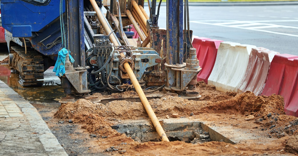 A blue directional drilling machine working beside traffic barriers with a pipe extending into a muddy street opening.