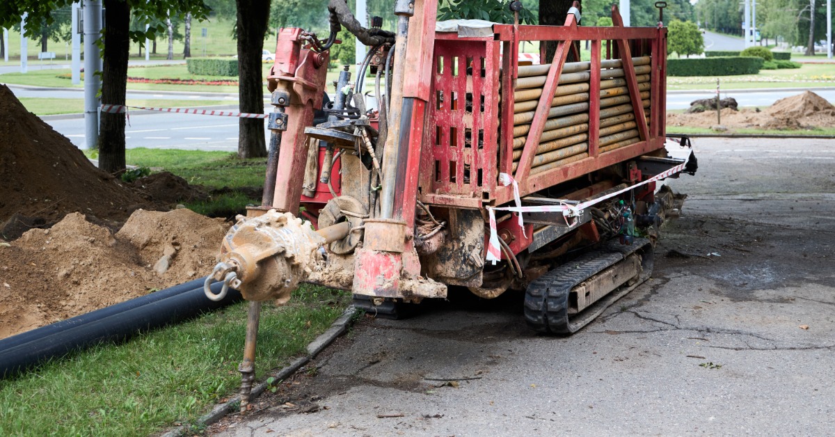 A red directional drilling machine parked on asphalt near trees, soil piles, and a black pipe along the ground.