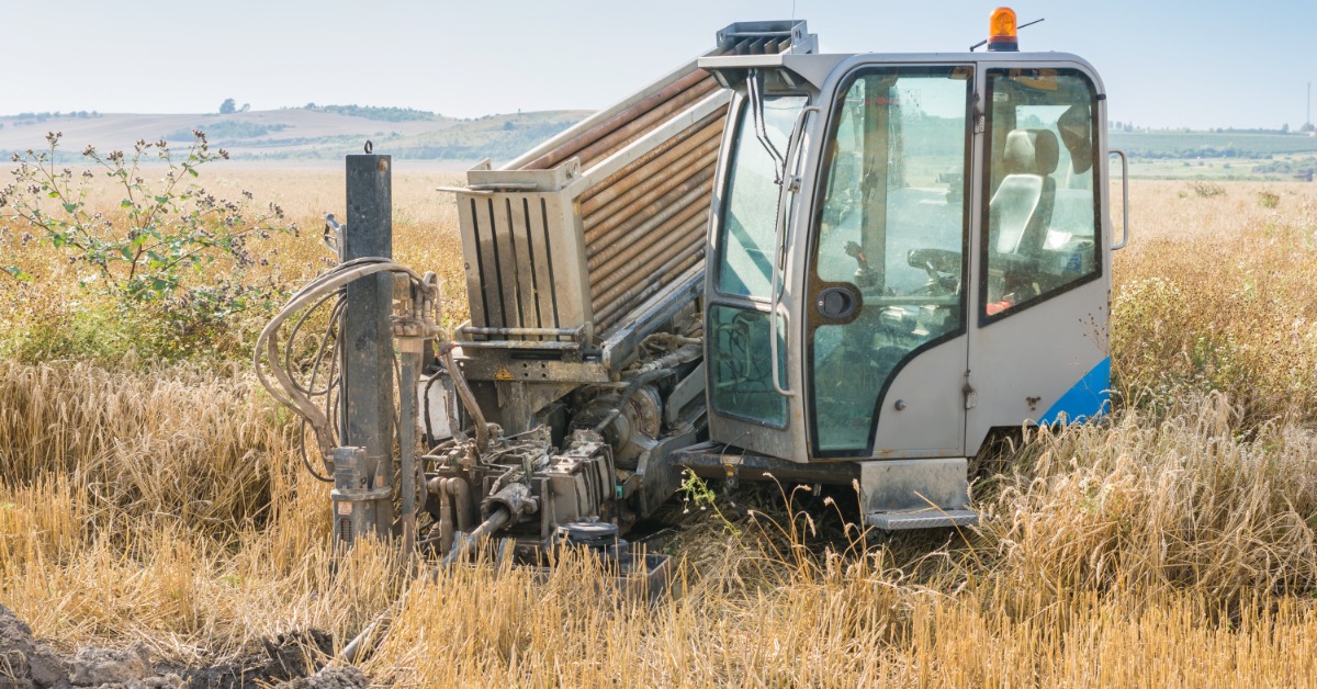 A white drilling machine with stacked rods parked in a dry field with tall grass and exposed soil in front.