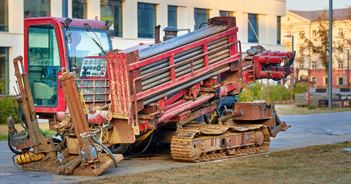 A red directional drilling machine parked on a paved walkway beside grass, benches, and modern buildings.