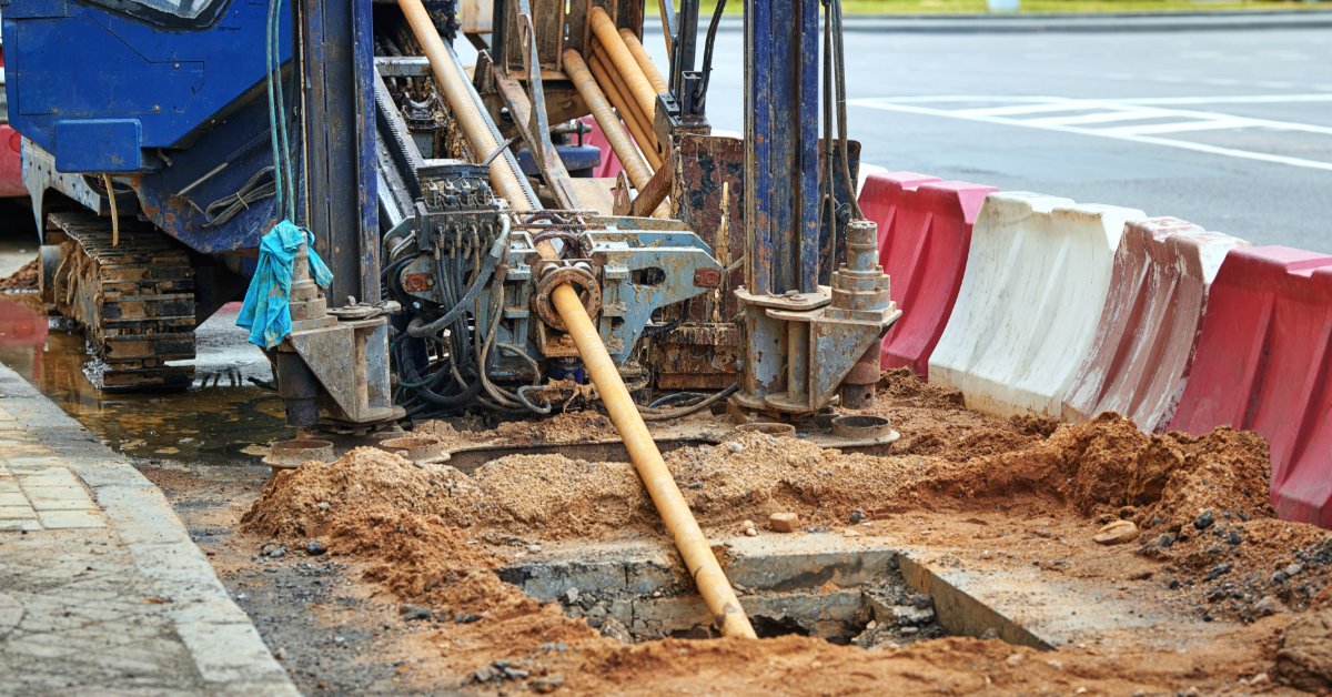 A blue drilling machine working beside red and white road barriers, with drill rods extending into a muddy trench.