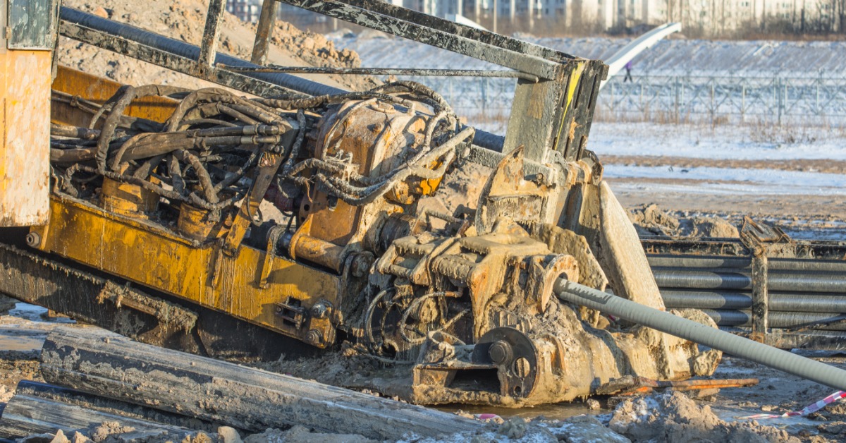 A muddy yellow directional drilling machine laying a gray pipe across a dirt construction area near buildings.