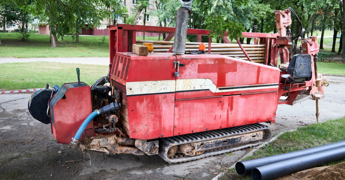 A red directional drilling machine parked on pavement near black pipes, grass, trees, and a dirt trench.