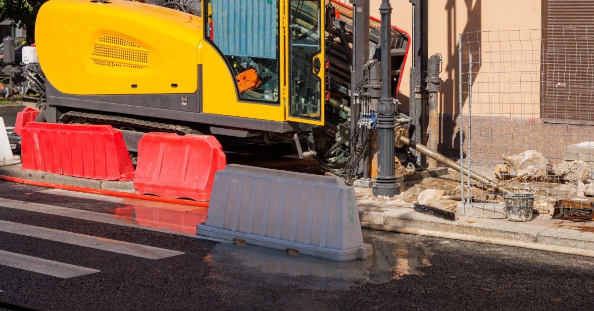 A yellow horizontal drilling rig operating beside traffic barriers on a city street with wet pavement.