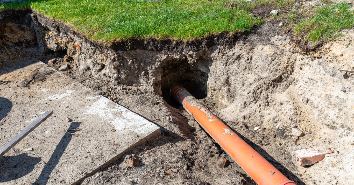 An orange pipe lying in an excavated trench beside grass, concrete, loose soil, and scattered debris.