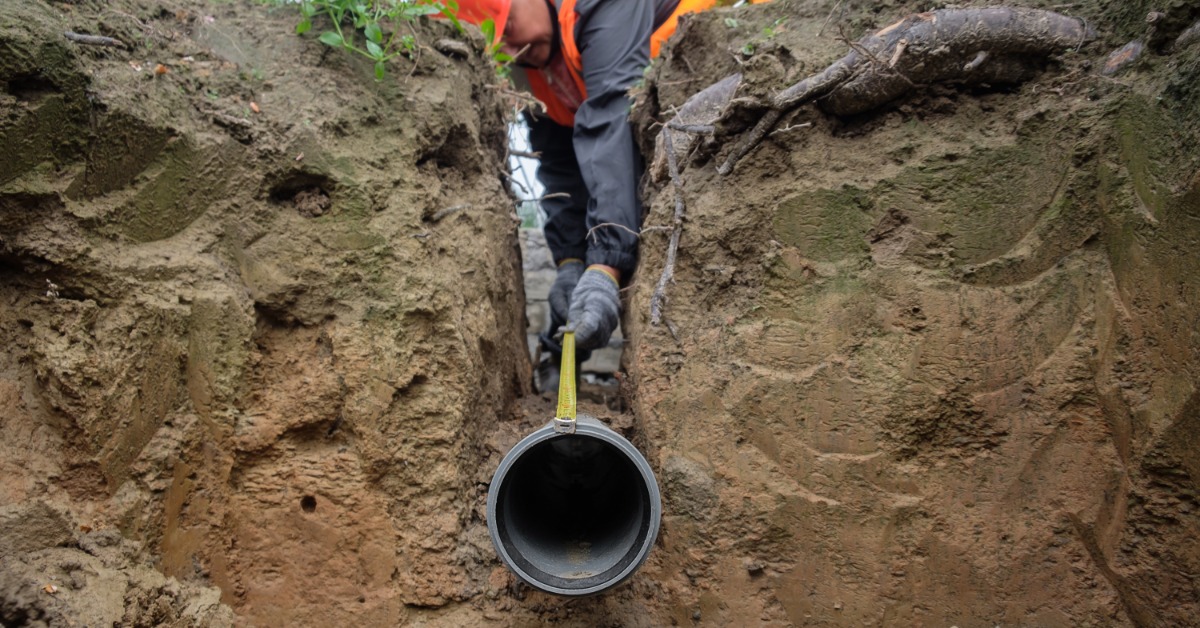 A person in a hard hat and orange vest measuring a black pipe inside a narrow excavated trench with exposed roots.