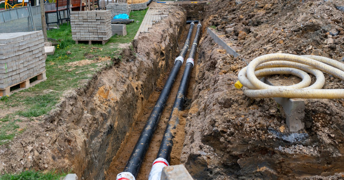 A long trench holding black pipes beside coiled conduit, stacked paving blocks, grass, and piles of excavated soil.
