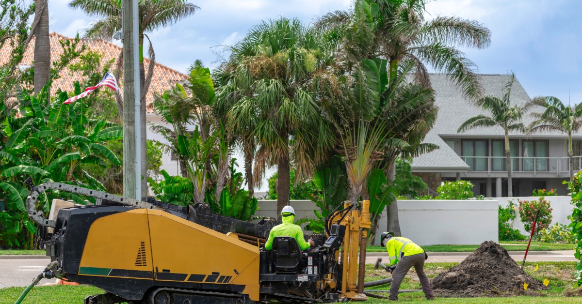 Horizontal directional drilling machine with two workers boring beside a palm-lined roadside near homes.