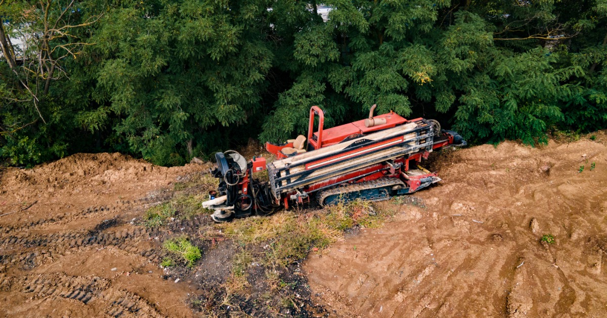 Red horizontal directional drilling machine on a dirt worksite beside dense trees, viewed from above.
