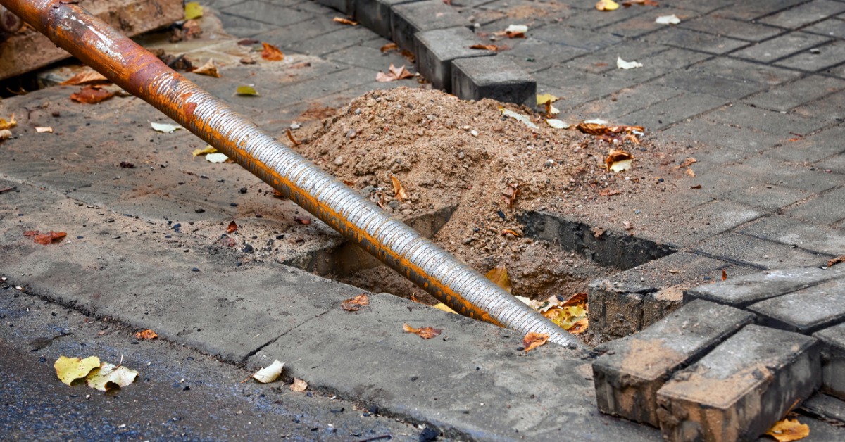 Directional drilling rod entering a small street excavation beside a paved sidewalk with loose soil and fallen leaves.