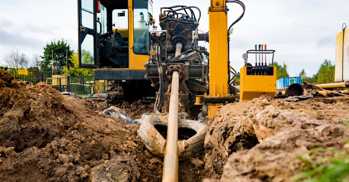 Horizontal drilling equipment guides a drill rod through an excavated city street trench filled with muddy water.