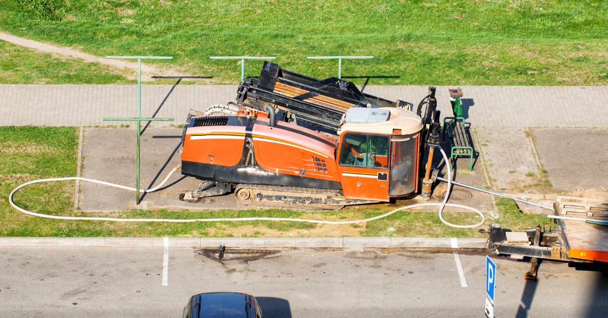 Orange horizontal directional drill sits beside a paved road, with hoses extending across grass near a parked vehicle.