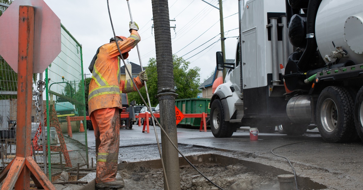 A worker in orange safety gear holding a hose near an excavation pit beside a white hydrovac truck on pavement.