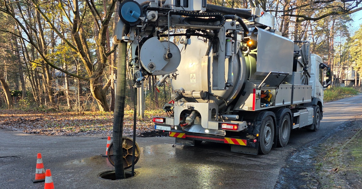 A white hydro-vac truck parked on a forest road with a large hose extending into an open manhole near cones.