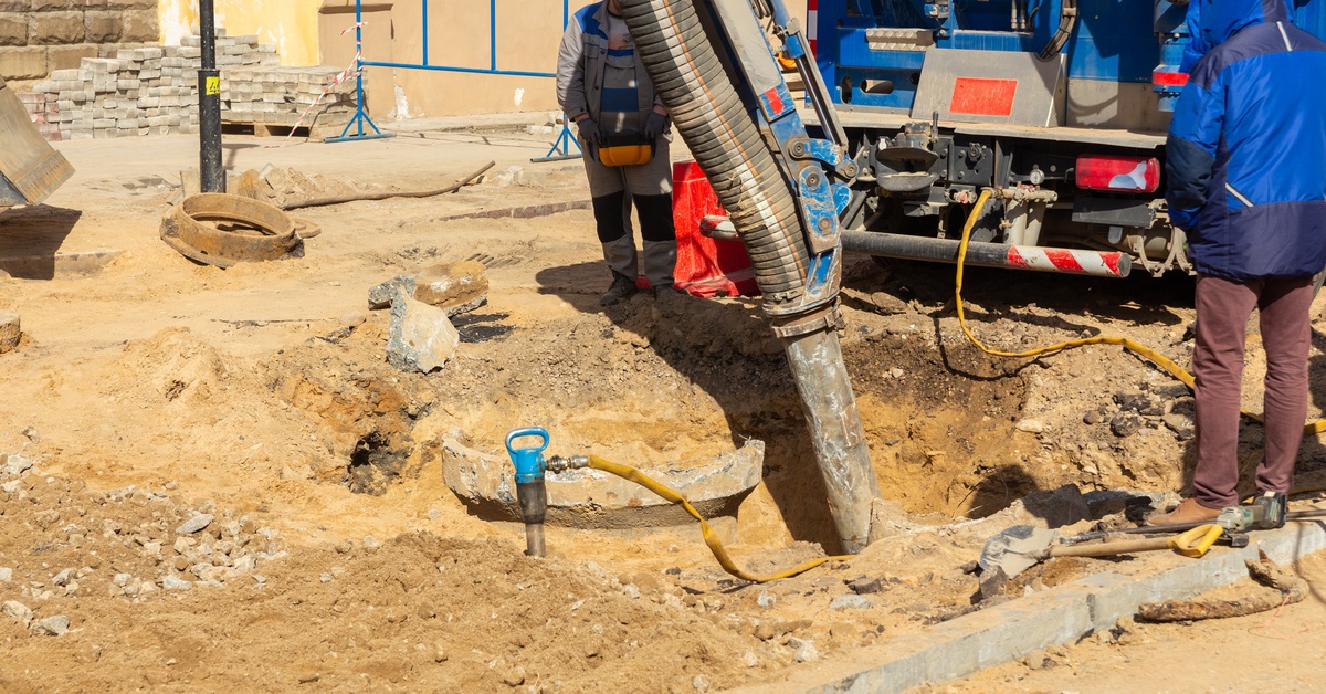 Two workers using a hose beside a suction excavator truck near an open utility hole on a paved road.