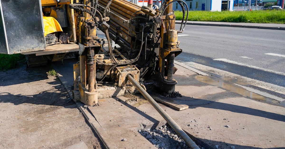 Directional drilling machine bores beneath a paved roadside while drill rods extend from the rig into the ground.