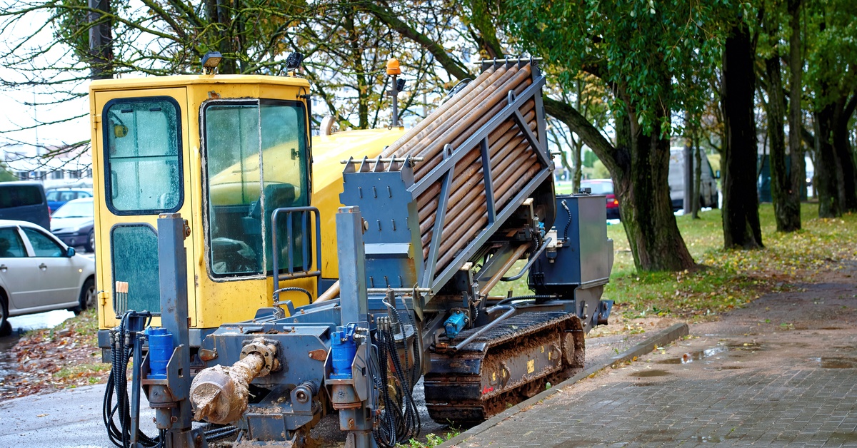 Yellow directional drilling machine with stacked drill rods sits beside a tree-lined sidewalk on a wet roadside.