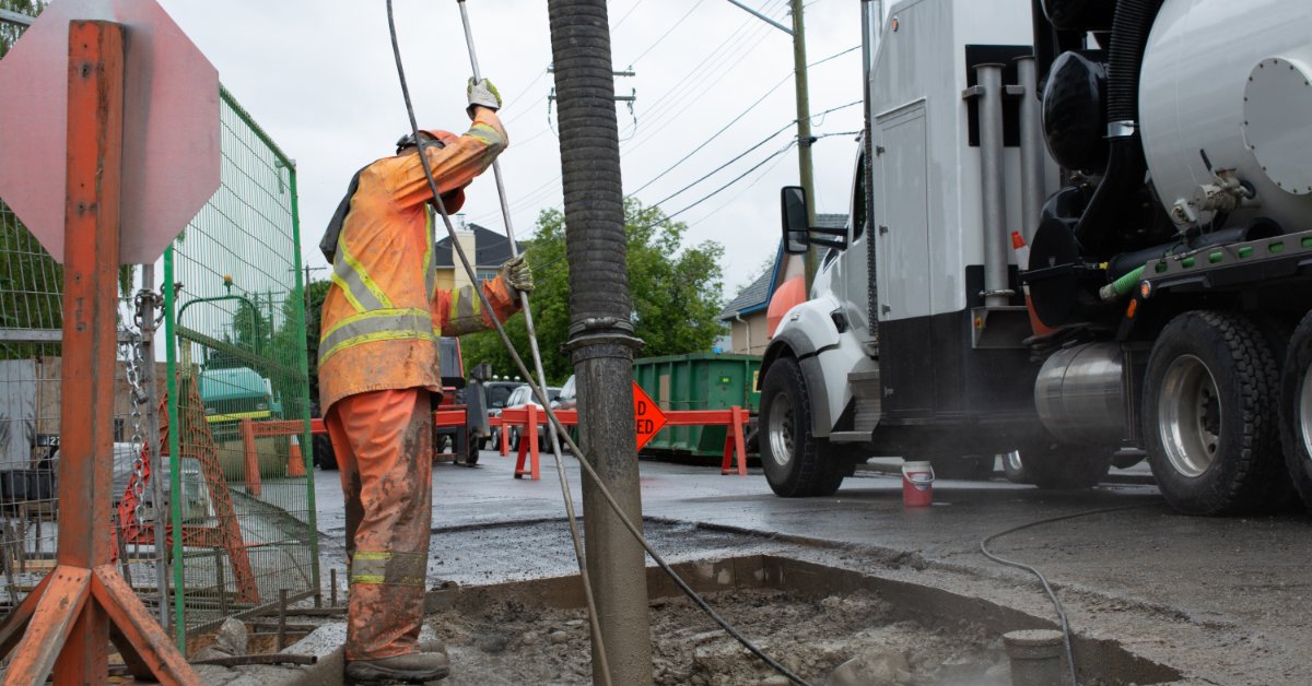 Worker uses a vacuum excavation hose beside a hydrovac truck to remove soil from a large roadside excavation.