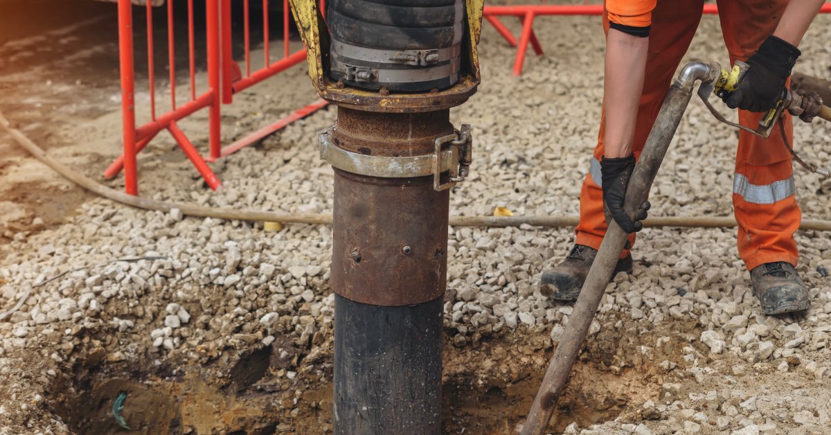 Worker uses a vacuum excavation wand beside a large suction hose to remove soil from a gravel-covered excavation.