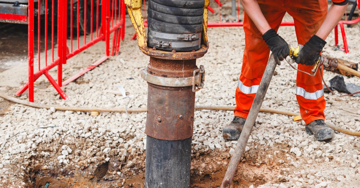 Worker uses a vacuum excavation wand beside a large suction hose to remove soil from a gravel-covered worksite.
