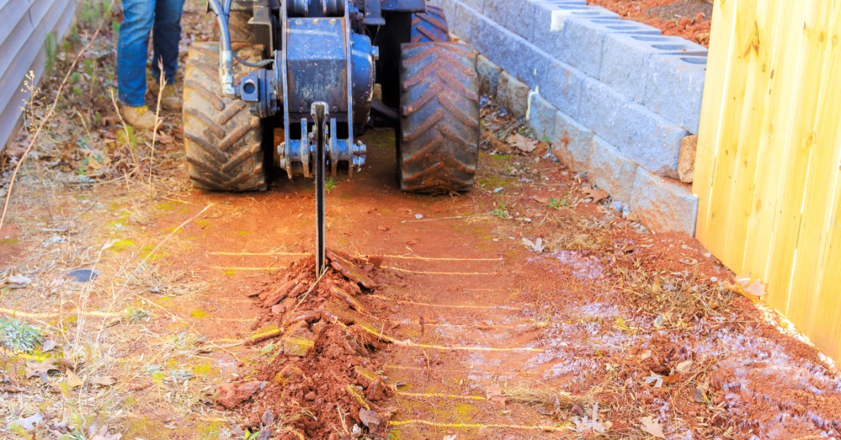 A cable plowing machine cuts a narrow trench through reddish soil beside a concrete block wall and yellow wooden fence.