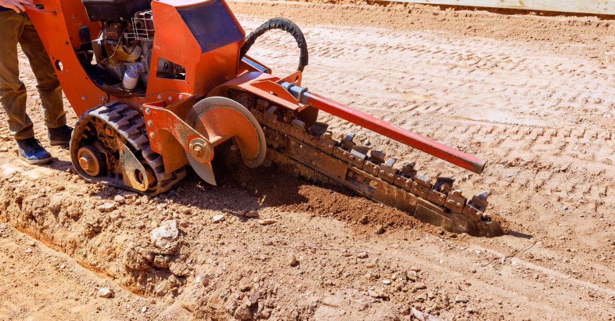 An orange tracked trencher cuts a narrow trench through dry, rocky soil, with a worker standing beside the machine.