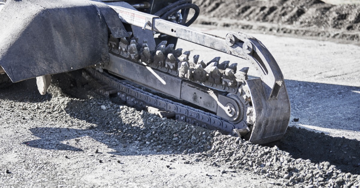 A trencher's metal digging chain cutting a narrow trench through rocky ground, with loose gravel piled alongside.
