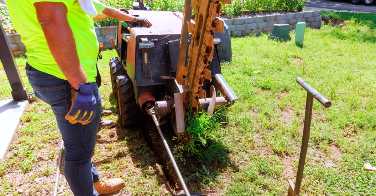 Worker in a neon yellow shirt operates trenching equipment, cutting a narrow trench through a grassy residential yard.